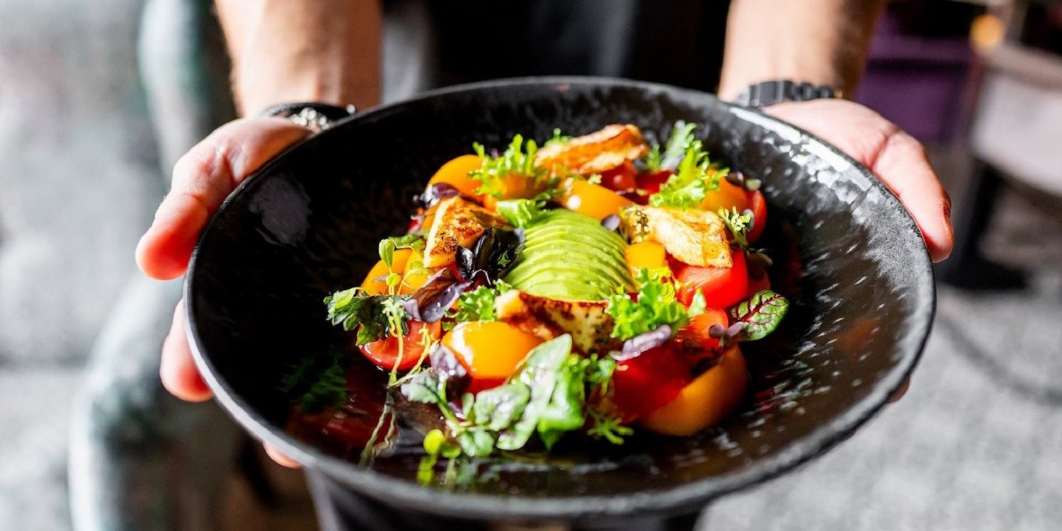 Eine Person hält einen schwarzen Teller mit einem bunten Salat aus Gemüse, Tomaten, Avocado und Tofu in der Hand. . Artikel "Mehr Pflanze auf den Teller bitte!" öffnen.
