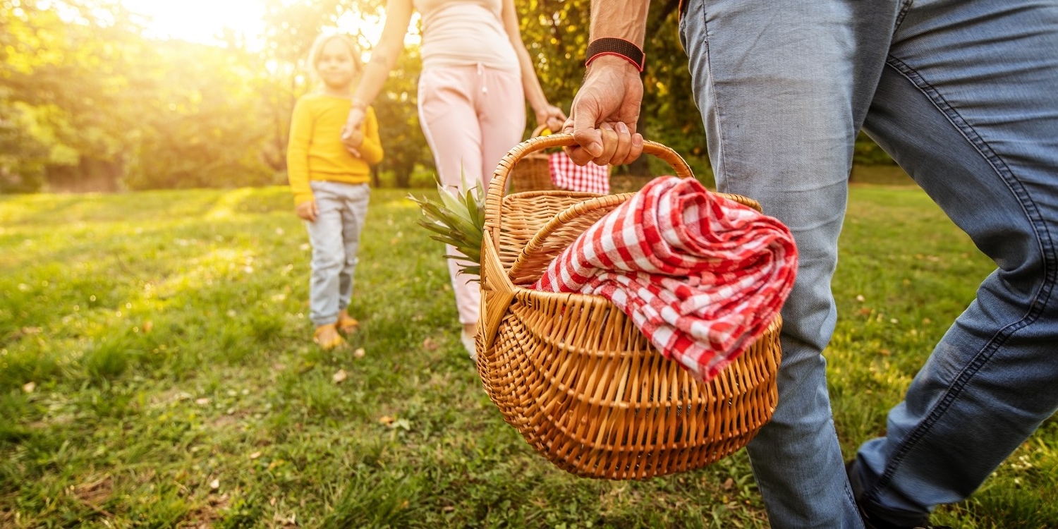 Ein Mann geht über eine Wiese und trägt einen Picknickkorb. Hinter ihm her laufen eine Frau und ein Kind. . Artikel "Genussmomente unter freiem Himmel " öffnen.