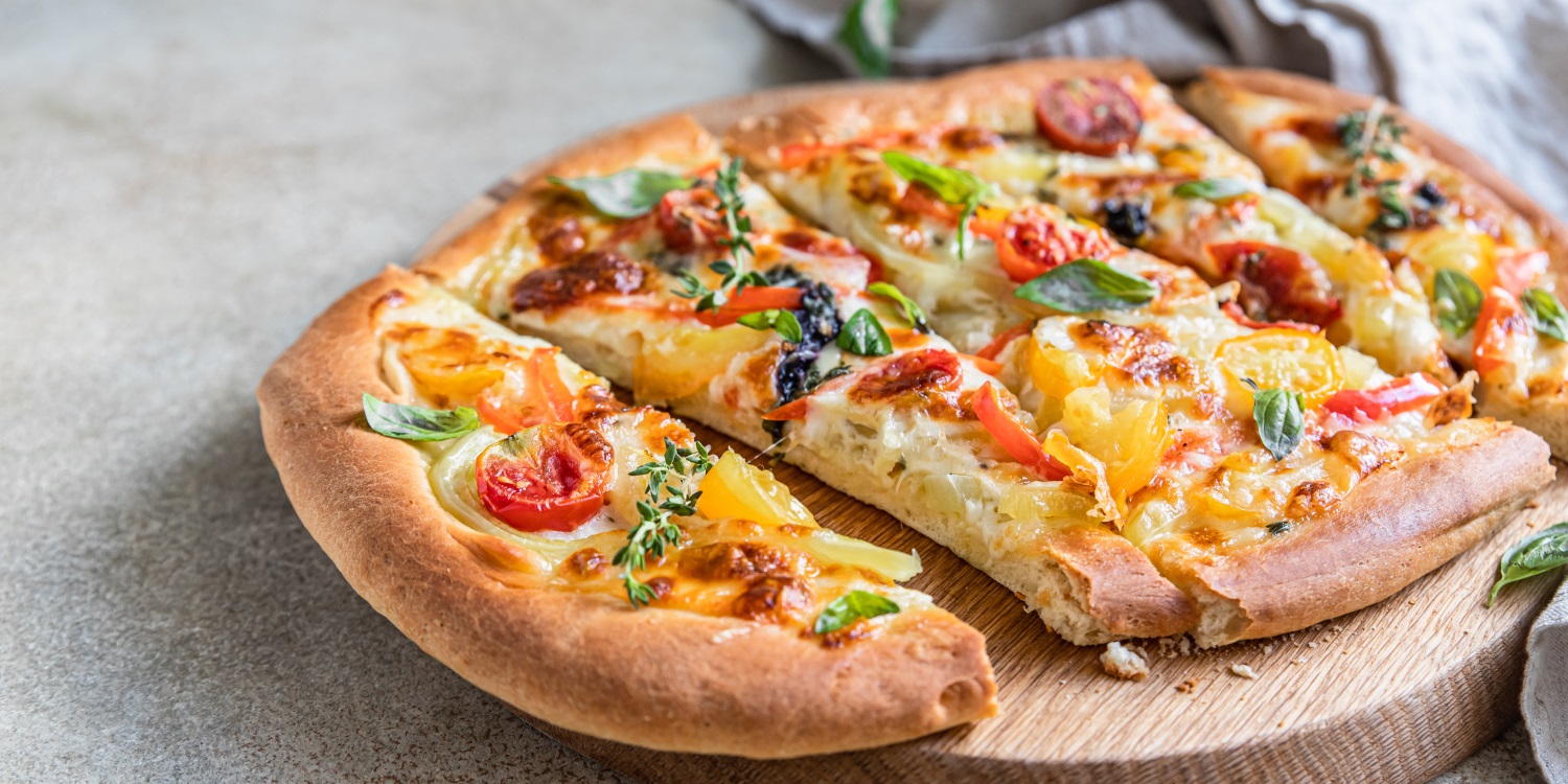 Auf einem Holzbrett liegt eine ovale Pinsa mit verschiedenfarbigen Tomatenstücken, Zwiebeln, Käse und Kräutern. . Artikel "Pinsa statt Pizza" öffnen.