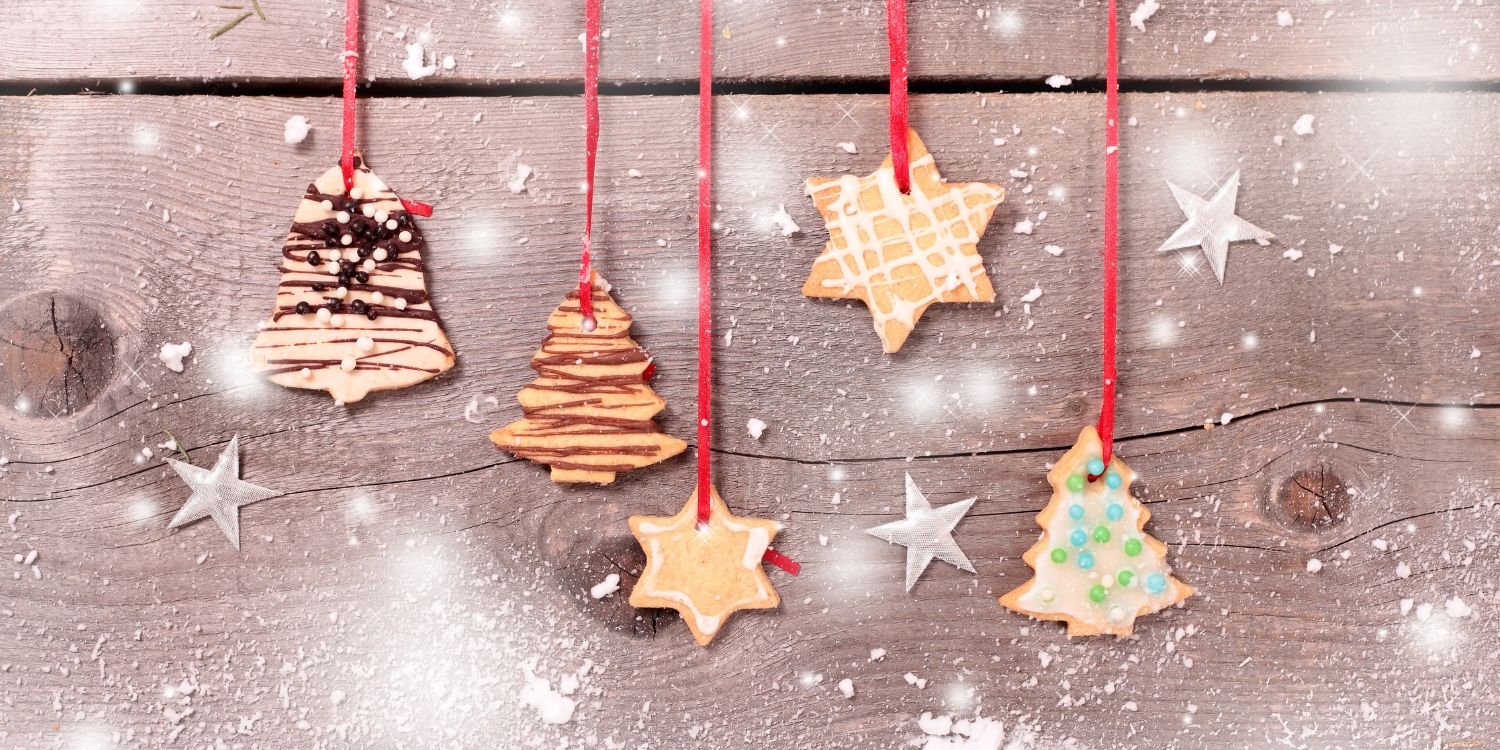 Vor einer grauen Holzverkleidung hängen verschieden geformte und dekorierte Plätzchen an roten Bändern, ringsum sind Schneeflocken.. Artikel "Christbaumschmuck zum Naschen" öffnen.