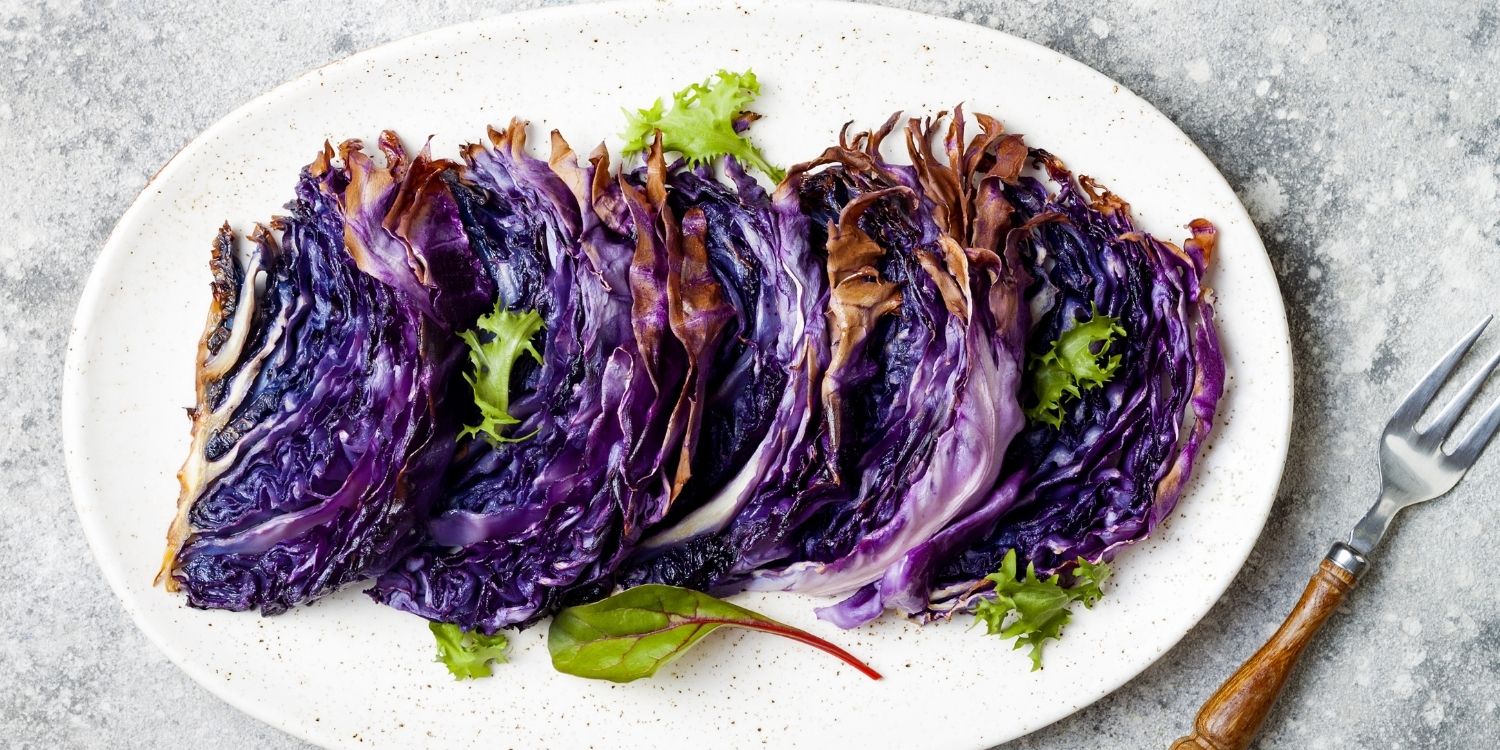 Auf einer ovalen weißen Platte liegen gegarte halbierte Rotkohlscheiben garniert mit Salatblättchen, daneben eine Gabel mit Holzgriff.. Artikel "Rotkohl kann auch anders" öffnen.