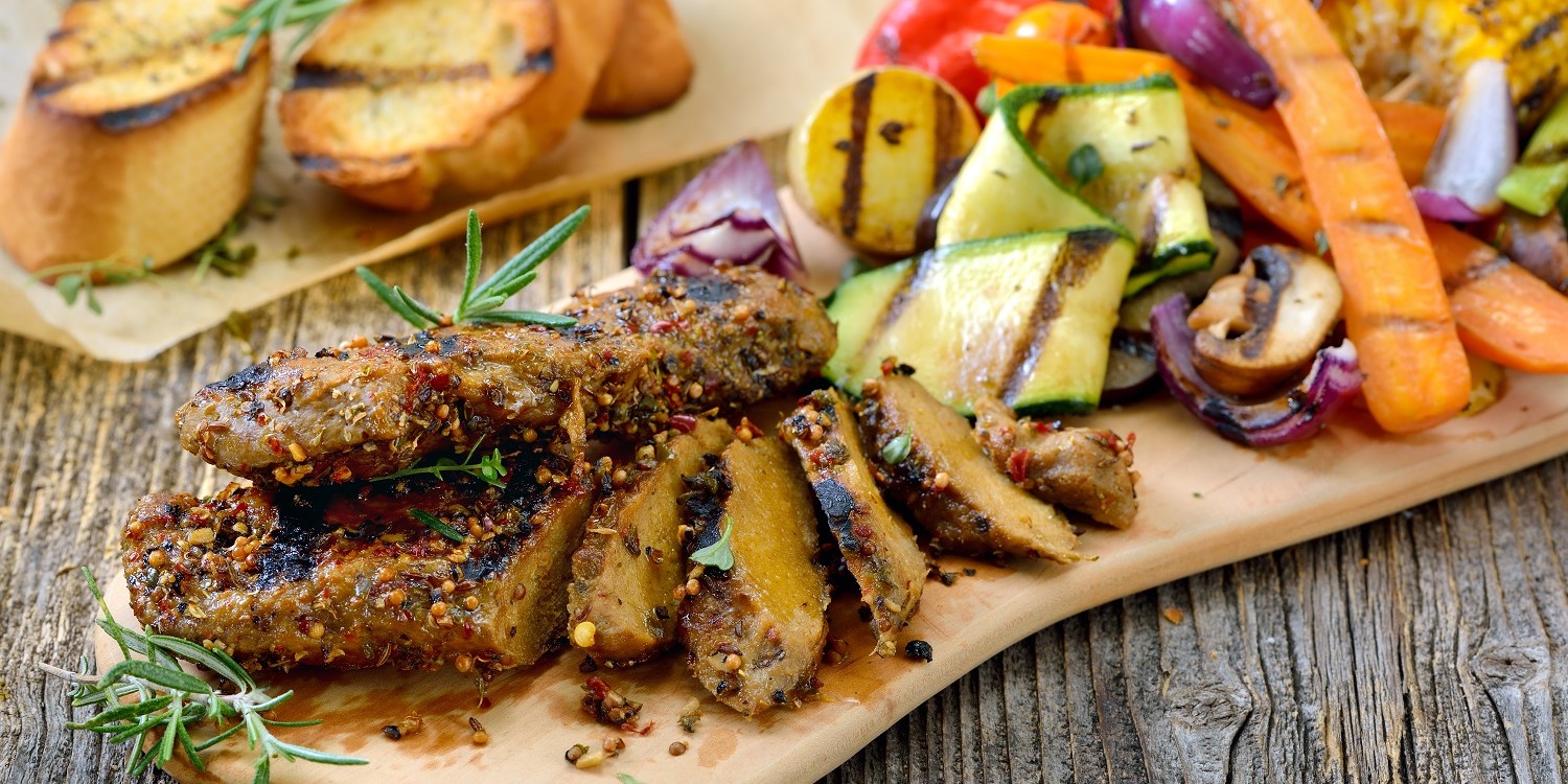 Auf einem Holzbrett liegt ein gegrilltes Seitan-Steak mit gemischtem Grillgemüse. Artikel "Alternativprodukte zu tierischen Lebensmitteln" öffnen.