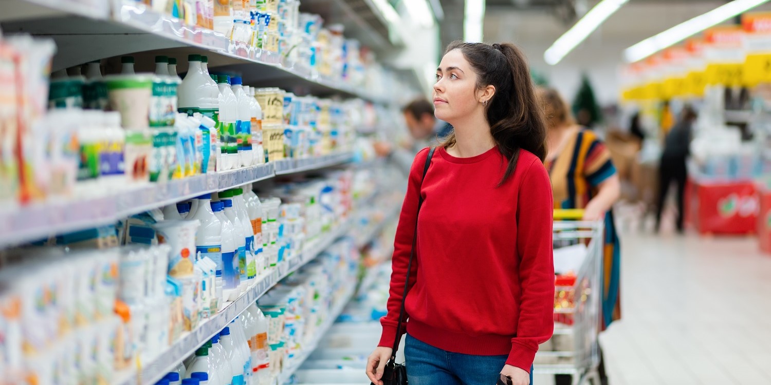 Eine junge Frau steht im Supermarkt vor einem Regal voller Lebensmittel. . Artikel "Was sind eigentlich hochverarbeitete Lebensmittel?" öffnen.