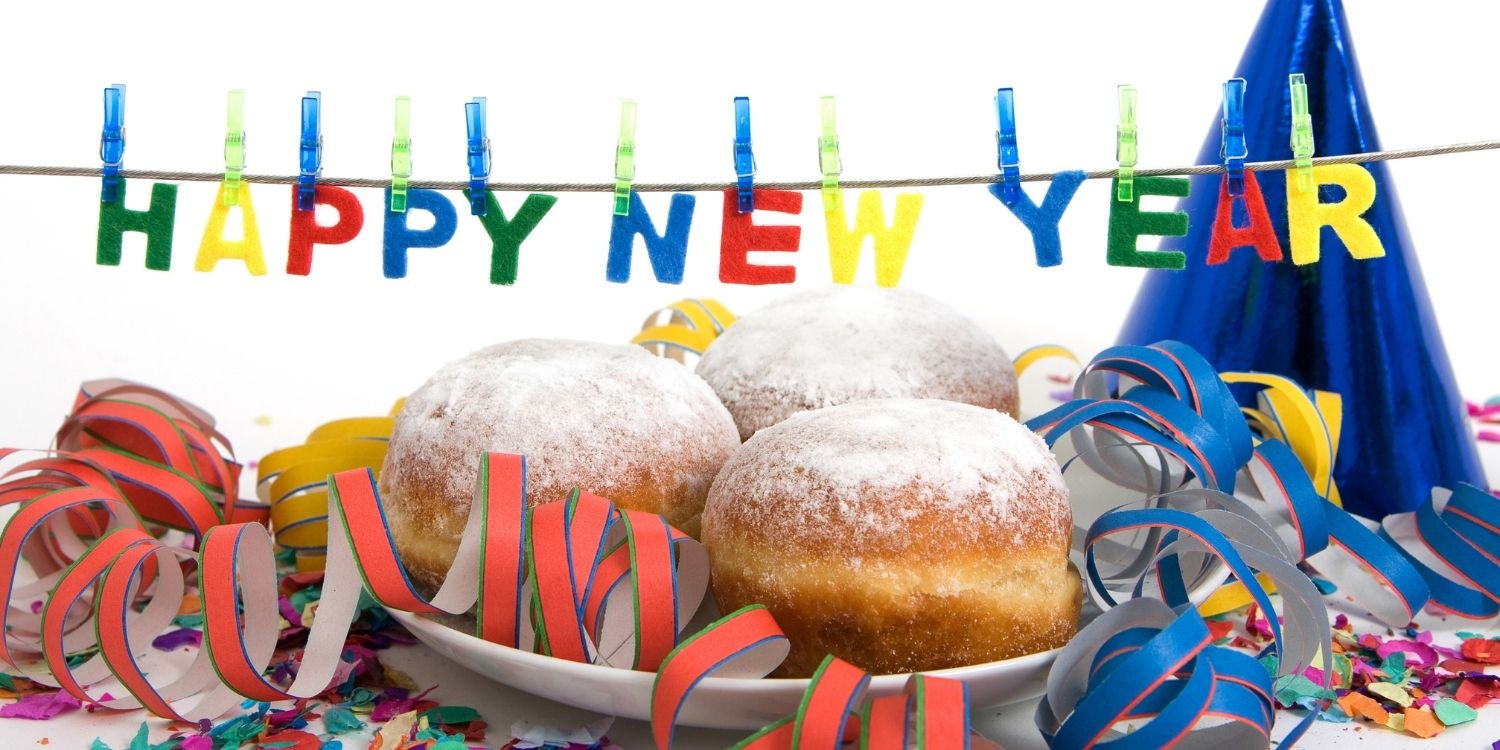 Auf einem Tisch steht ein Teller mit Berlinern, dekoriert mit Luftschlangen und darüber hängt eine Schnur mit Buchstaben an Wäscheklammern, die „Happy New Year“ formen.. Artikel "Kulinarische Neujahrsbräuche aus aller Welt" öffnen.