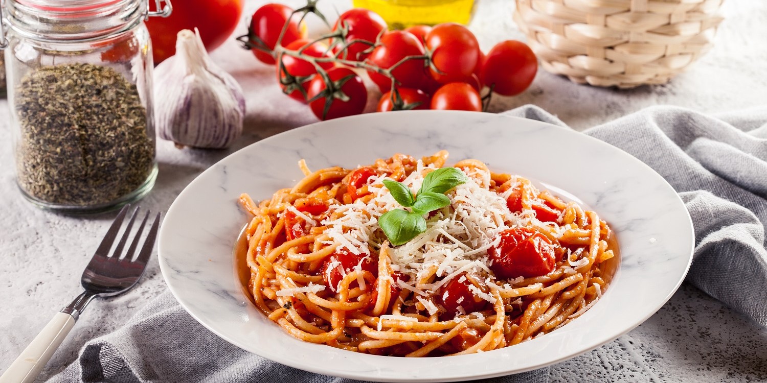 Auf einem weißen Teller befinden sich Spaghetti mit Tomatensoße und Parmesankäse. Hinter dem Teller steht ein Glas mit einer italienischen Kräutermischung, eine Knoblauchknolle, frische Tomaten, eine Karaffe mit Öl und eine Basilikum-Pflanze. . Artikel "Oma, App oder Kochbuch?" öffnen.