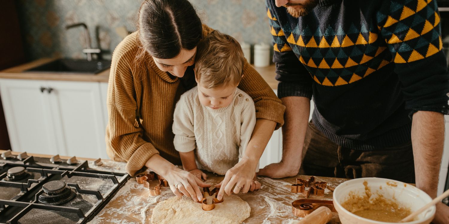 Eltern backen mit ihrem etwa zweijährigen Kind Plätzchen. Das Kind sticht mit einem Förmchen den ausgerollten Teig aus. . Artikel "Weihnachtsessen mit Kleinkindern" öffnen.