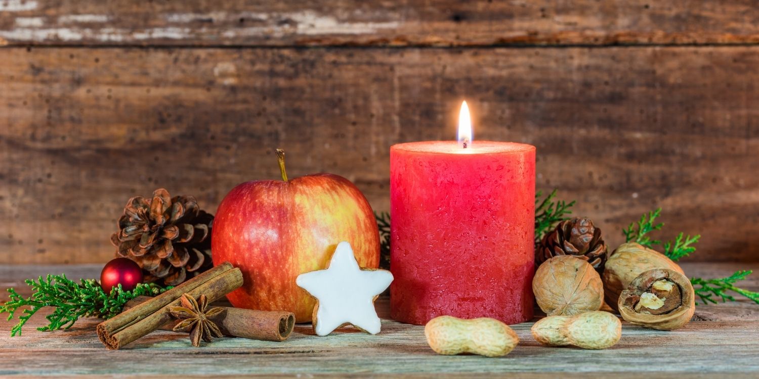 Vor einer Holzverkleidung ist eine brennende rote Kerze dekoriert mit Zweigen, Zapfen, Nüssen und Keksen. . Artikel "Weihnachtsgruß der Redaktion" öffnen.