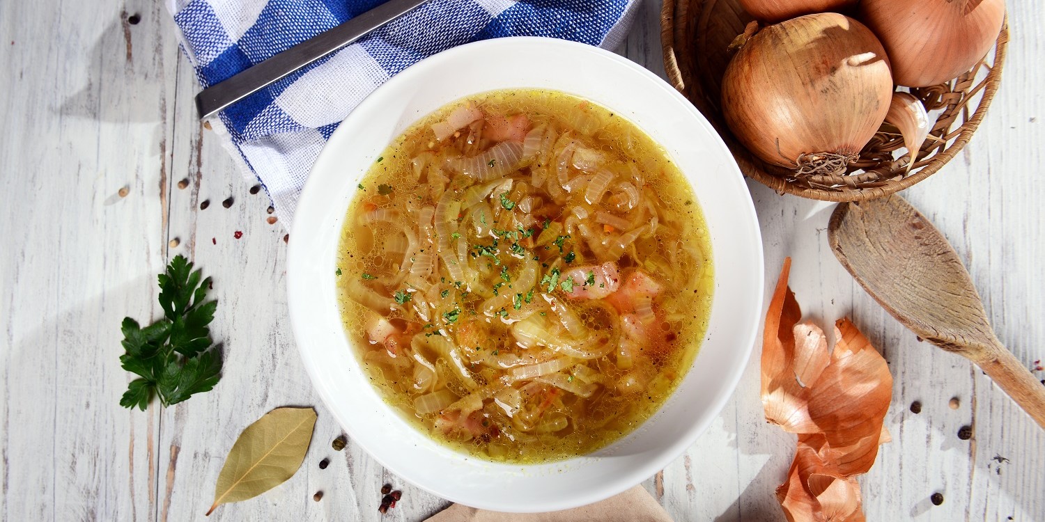 Selbstgemachte Zwiebelsuppe ist in einem weißen Teller angerichtet. Um den Teller herum liegen Petersilie, ein Lorbeerblatt, ein Kochlöffel und ein Bastkörbchen mit Zwiebeln. . Artikel "Ein Gemüse, das zu Tränen rührt" öffnen.