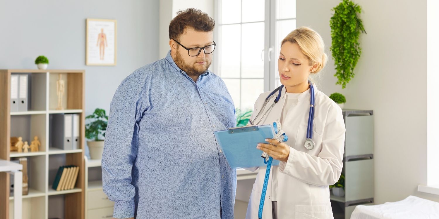 Ein kräftiger Mann im blauen Hemd und eine blonde Ärztin im Kittel schauen gemeinsam auf ein Klemmbrett.. Artikel "Neue Studie zu Adipositas-Kriterien" öffnen.