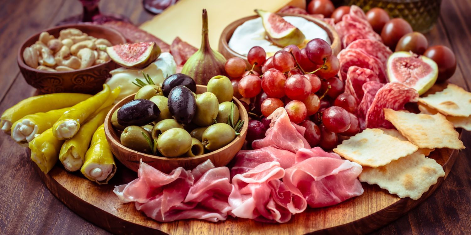 Auf einem runden Holzbrett sind Schinkenscheiben, Käse, Oliven, Trauben, Cracker, Feigen, gefüllte Peperoni und Nüsse angerichtet. . Artikel "Italienischer Antipasti-Guide" öffnen.