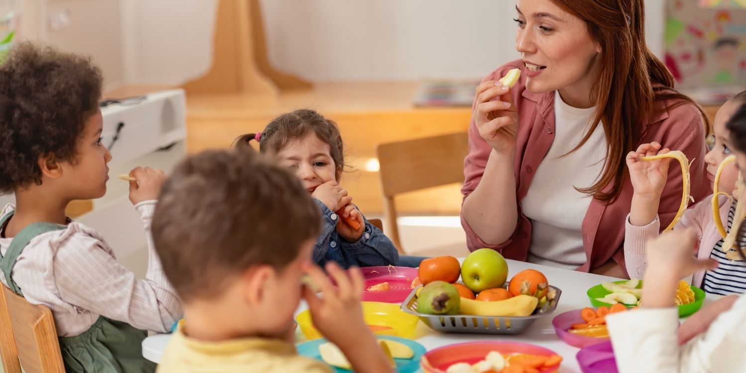 Eine Erzieherin sitzt mit drei Kita-Kindern an einem Tisch auf dem Obst und Gemüse in Stücke geschnitten steht und sie essen davon.. Artikel "Leckeres Essen in Kita und Kindertagespflege" öffnen.