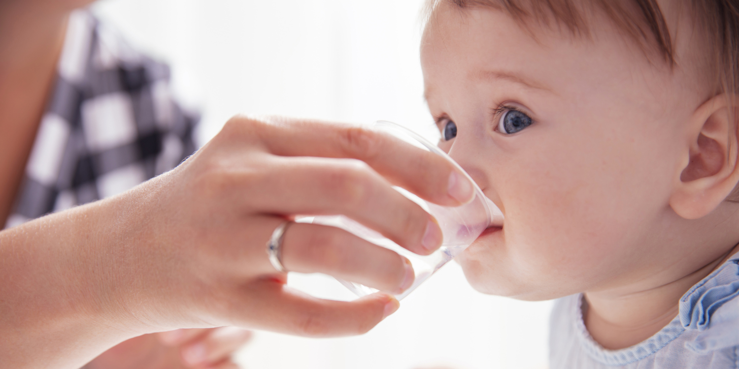  Eine Frauenhand hält einem Baby ein kleines Glas Wasser vor den Mund und es versucht zu trinken.. Artikel "Sollen Babys und Kleinkinder bei Sommerhitze mehr trinken?" öffnen.