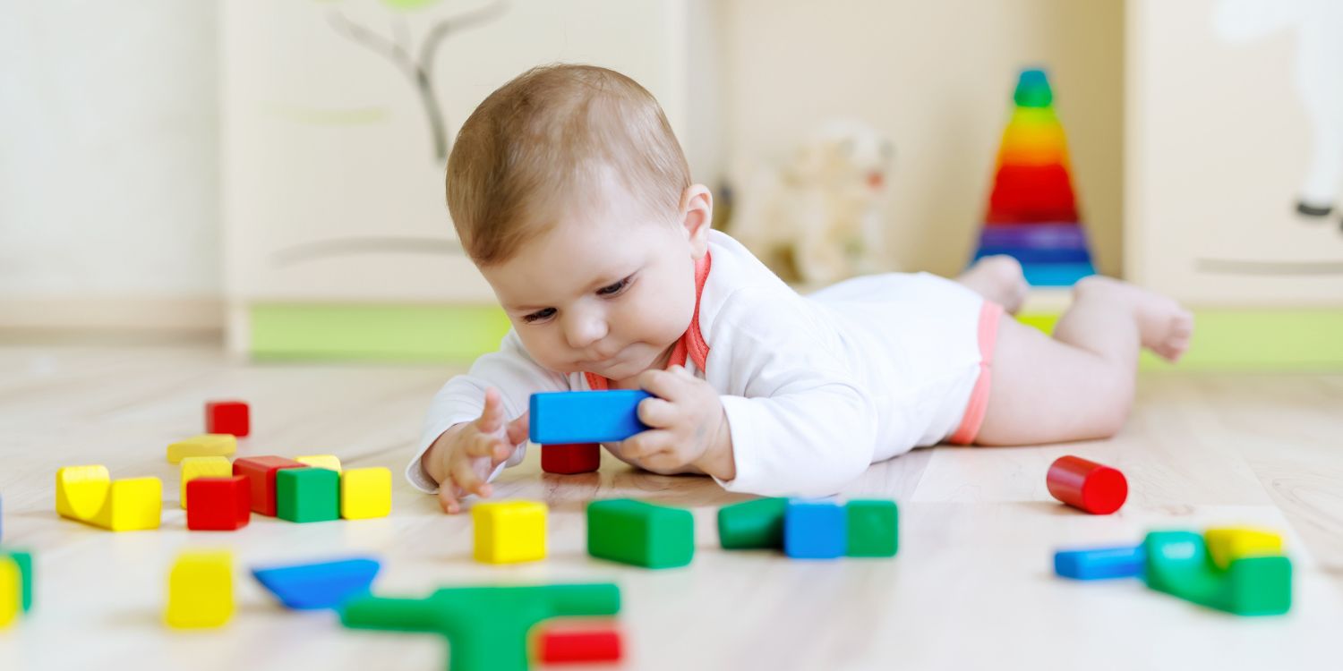 Ein Baby liegt auf dem Bauch auf dem Fußboden und spielt mit bunten Bauklötzen. . Artikel "Bewegung für die Kleinsten" öffnen.