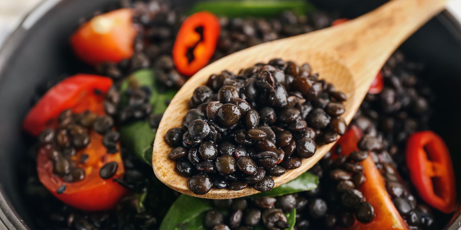 Gekochte Beluga-Linsen auf einem Holzlöffel über einer Schüssel mit einem Linsen-Salat mit Tomaten, Spinat und Chilis. . Artikel "Welche Linse für welches Gericht?" öffnen.