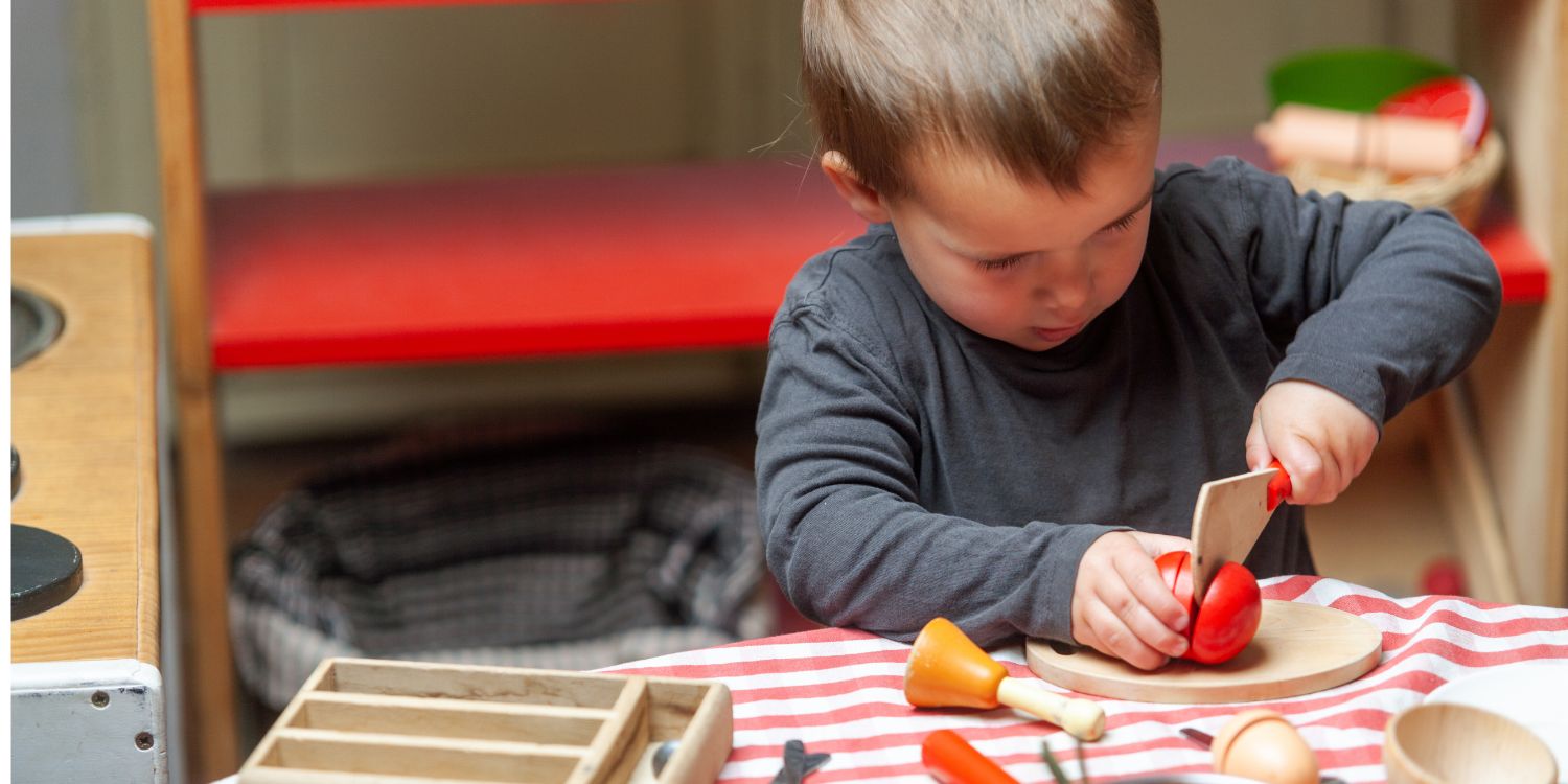 Ein Junge schneidet in einer Spielküche konzentriert Holztomaten mit einem Holzmesser.. Artikel "Ernährungsbildung in Kita und Kindertagespflege" öffnen.