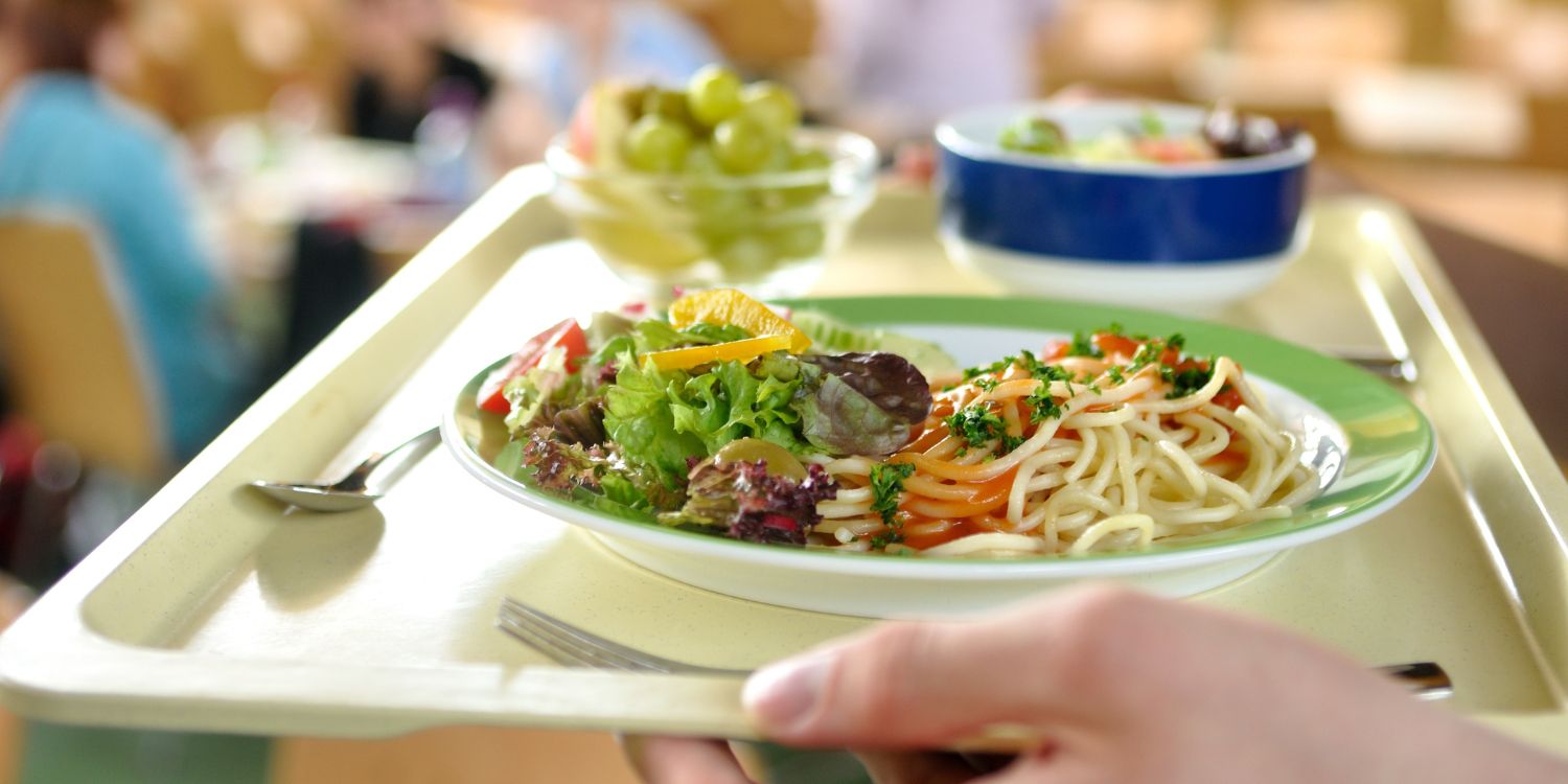 Hände tragen ein Tablett mit Essen in der Schulmensa. Auf einem Teller sind Nudeln mit Sauce und Salat, daneben seht eine Glasschale mit Trauben und eine Schale mit Gemüse. . Artikel "Bundeskongress Kita- und Schulverpflegung 2026" öffnen.