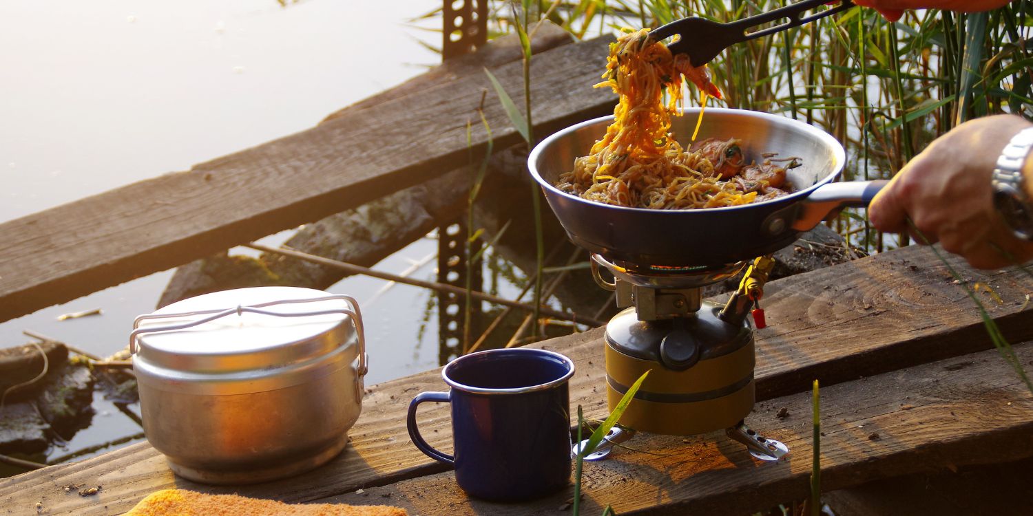 Eine Person kocht auf einem Campingkocher ein Nudelgericht vor einem See.. Artikel "Gut essen im Campingurlaub" öffnen.