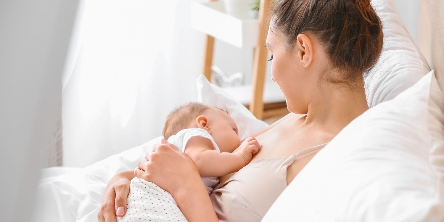 Eine junge Frau sitzt zuhause im Bett und stillt einen Säugling.. Artikel "„Clusterfeeding“ – Stillen nach Bedarf " öffnen.