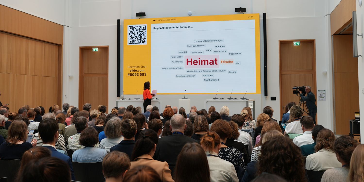 Der mit Besucherinnen und Besuchern gefüllte Veranstaltungsraum des Deutschen Ernährungstags 2026. Im Vordergrund spricht eine Referentin. Auf der Leinwand ist eine Wortwolke, in der Mitte steht das Wort Heimat.  . Artikel "Regionale Ernährung in Aktion" öffnen.