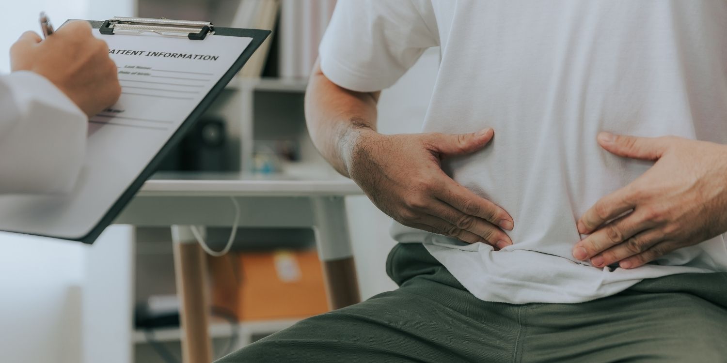 Ein Mann sitzt in einem Arztzimmer und hält sich mit beiden Händen den Unterbauch. Der Arzt neben ihm macht sich Notizen auf einem Klemmbrett.. Artikel "Tipps für die „Darmgesundheit“" öffnen.