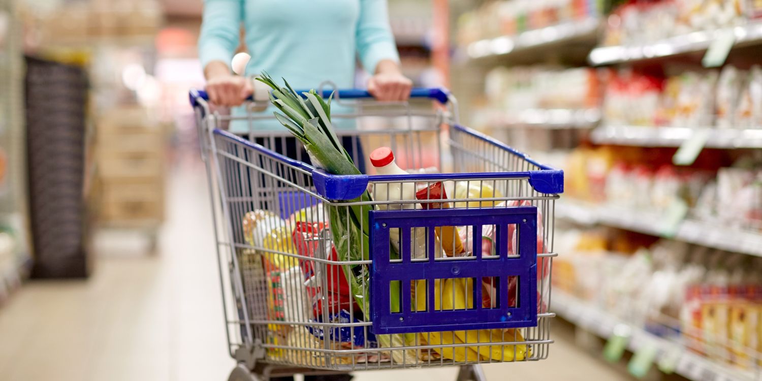 Eine Frau schiebt einen Einkaufswagen durch einen Supermarkt. Darin liegen Milch, Obst, Gemüse, Öl und Teigwaren.. Artikel "Zahlen zum deutschen Einkaufskorb" öffnen.