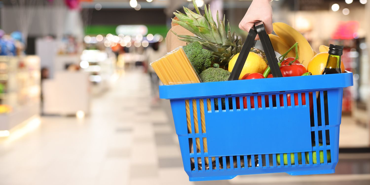 Eine Person im Supermarkt hält einen blauen Einkaufskorb mit Obst, Gemüse, Nudeln und Öl. . Artikel "Was beim Lebensmitteleinkauf zählt" öffnen.