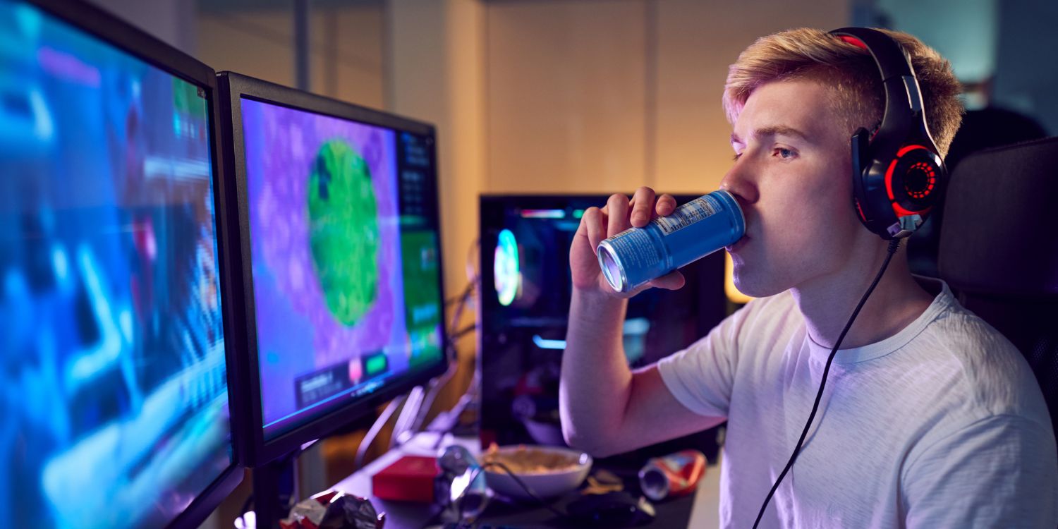 Ein Jugendlicher sitzt mit Kopfhörern vor mehreren Bildschirmen, auf denen man ein Computerspiel sieht, und trinkt aus einer Energydrink-Dose.. Artikel "Energydrinks im Deutschunterricht" öffnen.