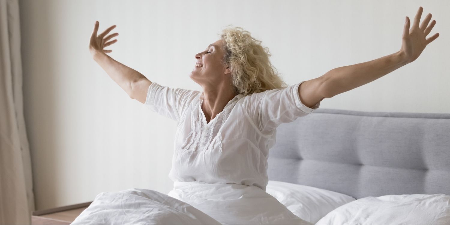 Eine ältere Frau sitzt im Bett und streckt genüsslich die Arme zu beiden Seiten aus.. Artikel "Gesunder Dreiklang: Ernährung, Schlaf, Bewegung" öffnen.