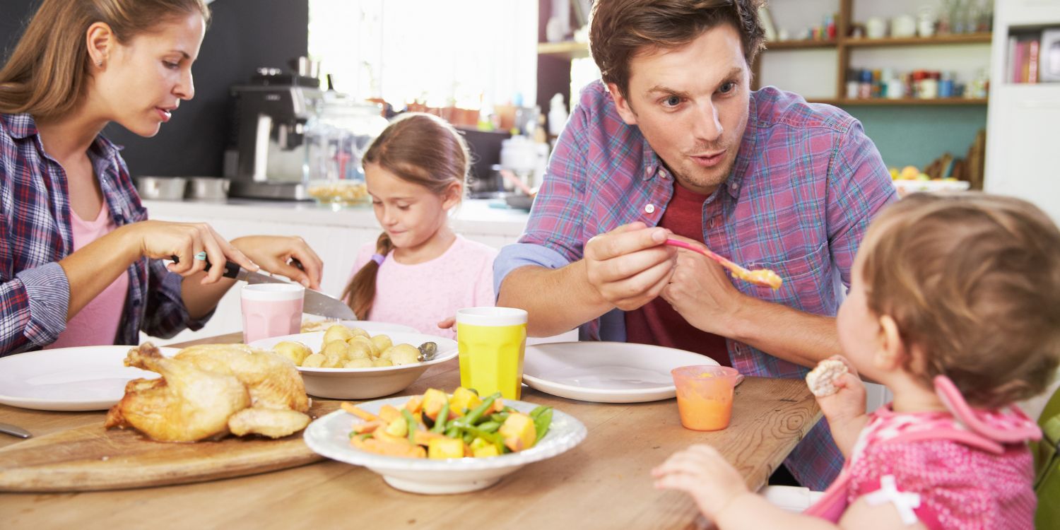 Ein Elternpaar sitzt in der Küche mit zwei Kindern beim Essen. Der Vater füttert das Kleinkind und die Mutter schneidet dem größeren Kind das Essen klein.. Artikel "Gemeinsam essen mit Kleinkindern " öffnen.