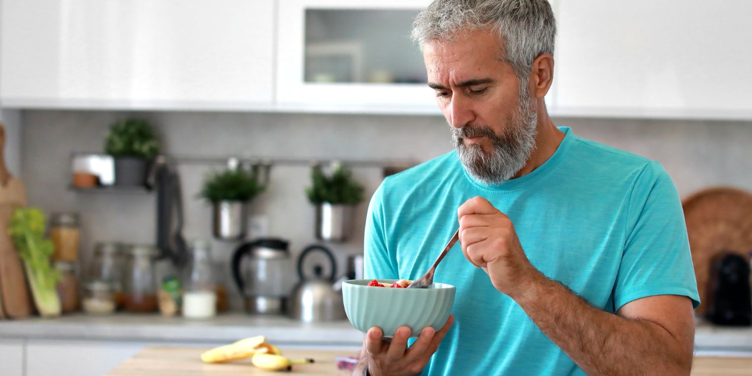 In einer Küche sitzt ein grauhaariger, bärtiger Mann im T-Shirt und isst Obst aus einer Schüssel. . Artikel "Gesund essen, langsamer altern?" öffnen.