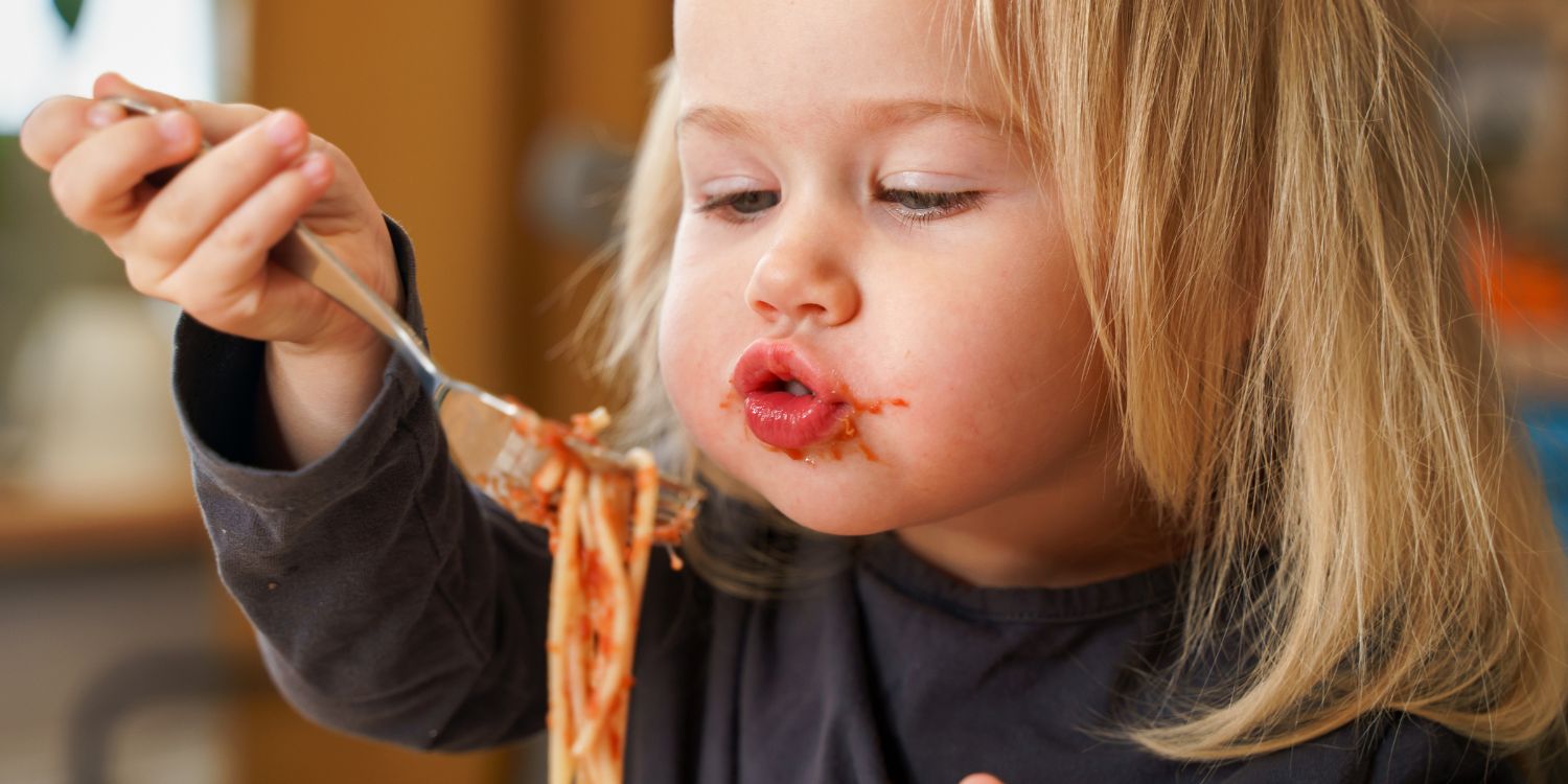 Ein blondes Mädchen im dunklen Oberteil führt mit einer Gabel etwas ungelenk Spaghetti mit Tomatensauce zum Mund und spitzt die Lippen. Ihr Mund ist mit Sauce verschmiert.. Artikel "Jedes Kind is(s)t einzigartig" öffnen.
