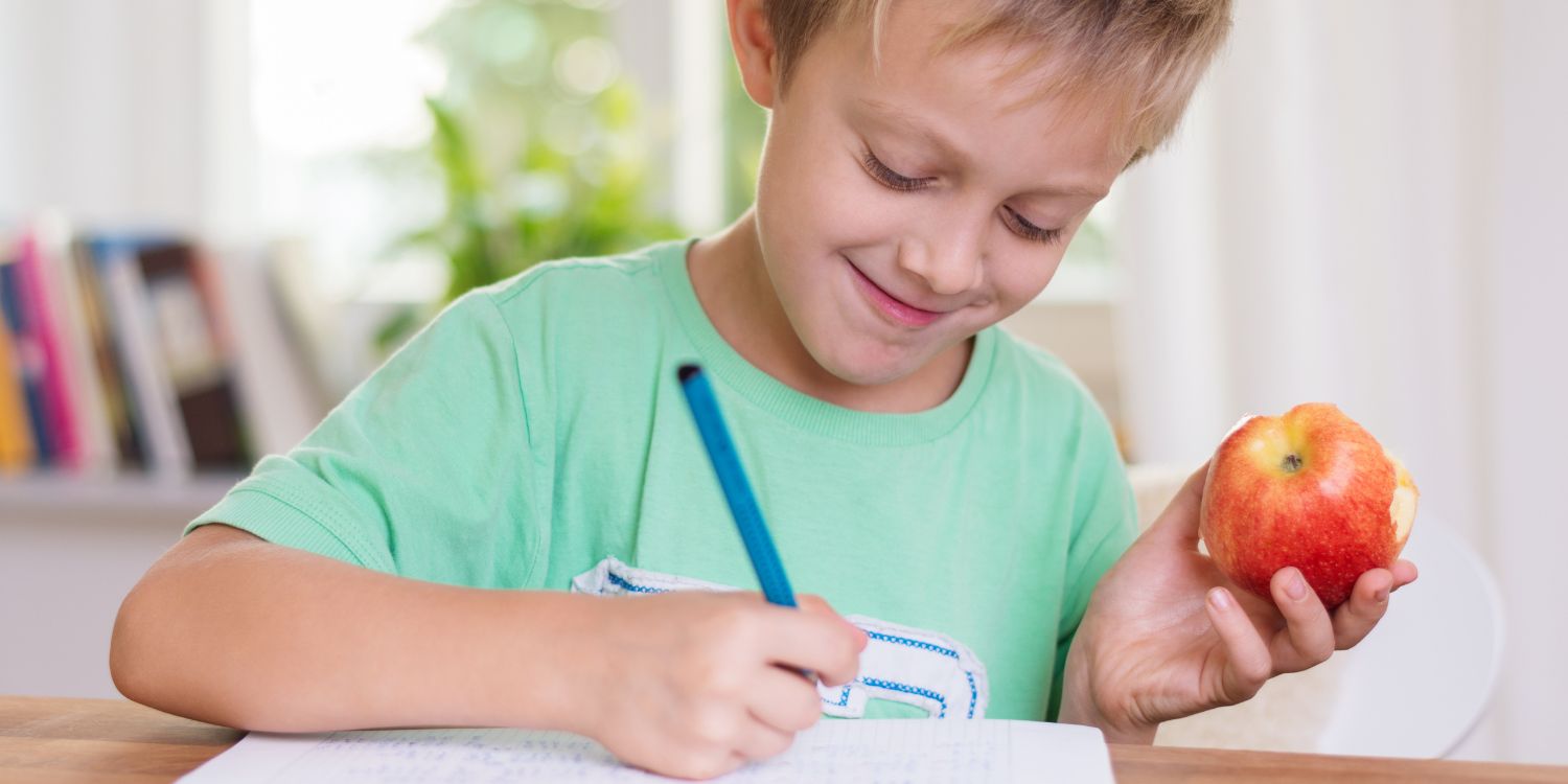 Ein blonder Junge im Grundschulalter sitzt an einem Tisch und schreibt in ein Heft. In der Hand hält er einen angebissenen Apfel.. Artikel "Der Feinschmeckerkurs" öffnen.