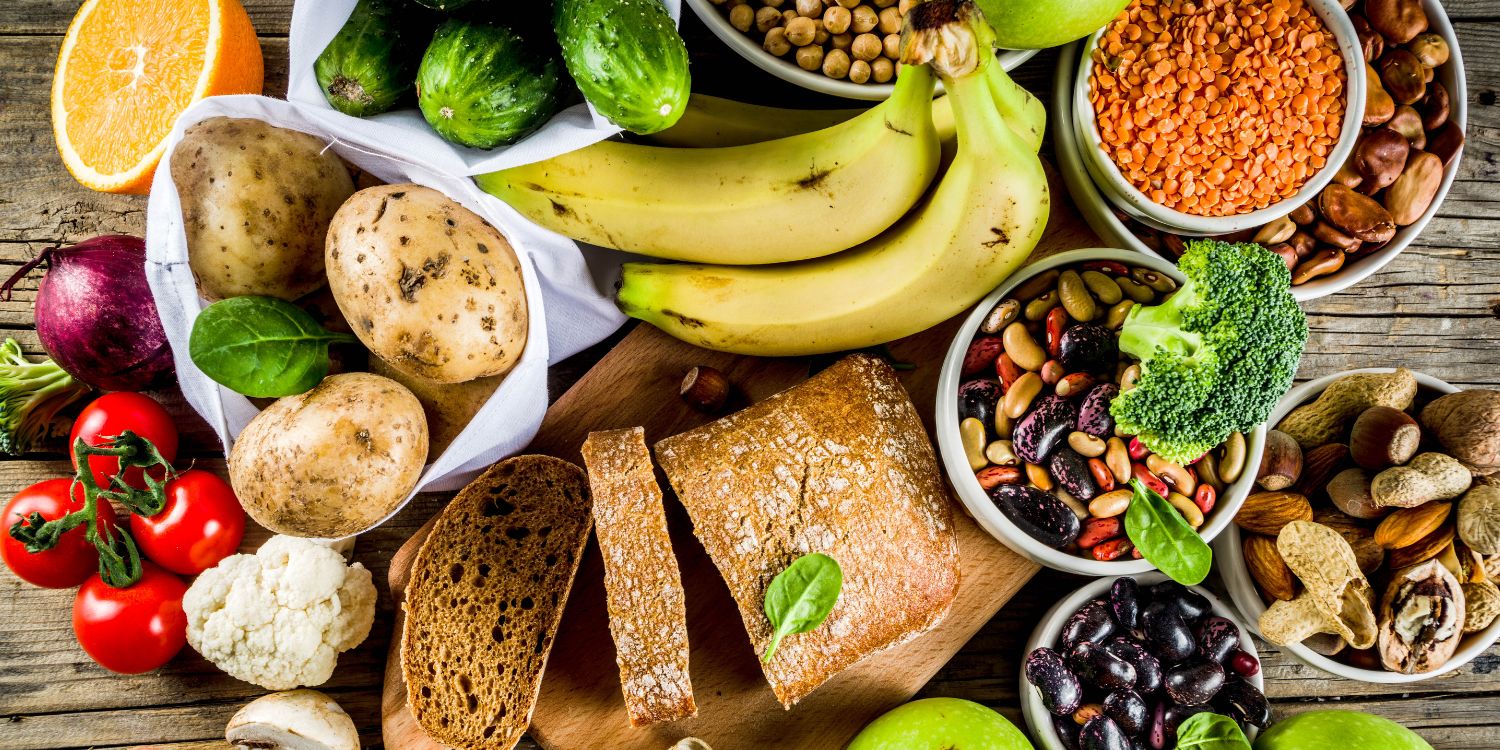 Auf einem Holztisch liegen viele ballaststoffreiche Lebensmittel wie Brot, Bananen, Kartoffeln, Linsen, Bohnen, Nüsse, Gemüse und Obst.. Artikel "Trend Fibermaxxing" öffnen.