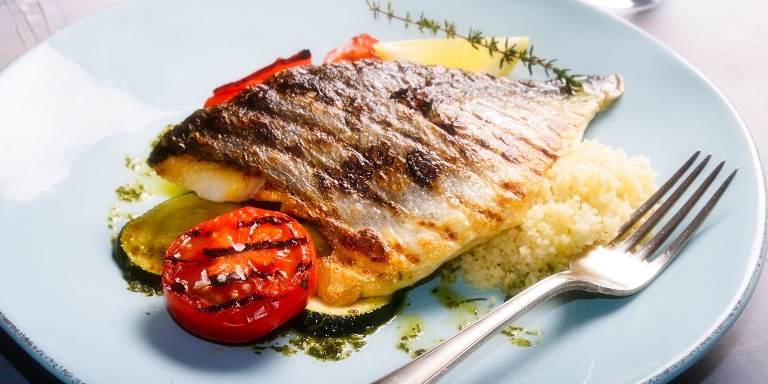 Auf einem weißen Teller liegt ein Stück gegarter Fisch garniert mit Zucchini, Tomate, Kräutern und Couscous neben einer Gabel.. Artikel "Fisch und Meeresfrüchte " öffnen.