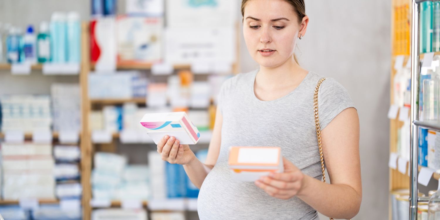Eine schwangere Frau steht in der Apotheke und hält zwei Verpackungen in den Händen. . Artikel "Folsäure – wichtig vor und während der Schwangerschaft" öffnen.
