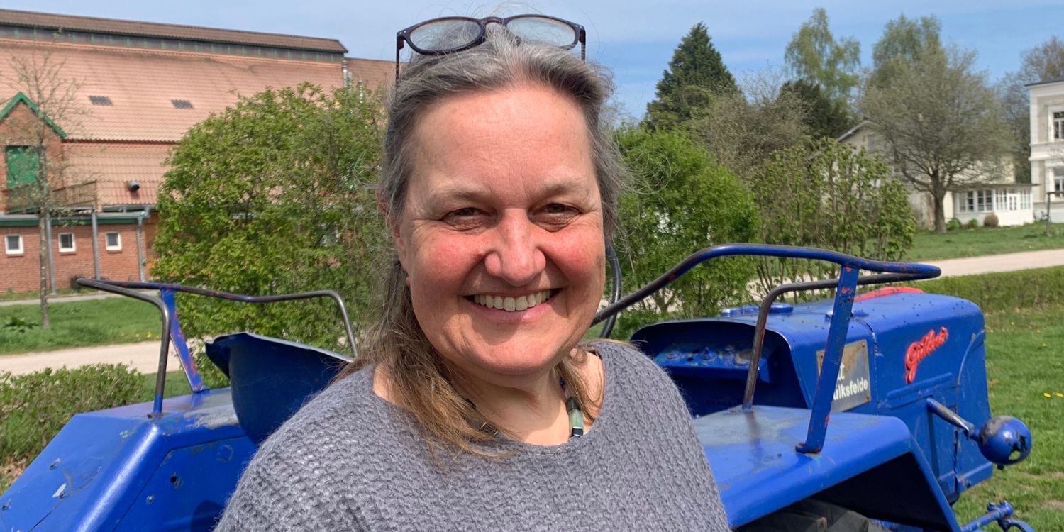 Eine dunkelhaarige Frau mittleren Alters steht im Freien vor einem Traktor und schaut in die Kamera, im Hintergrund sind landwirtschaftliche Gebäude.. Artikel "#11 Mit Körnern, Kindern und Kantinen" öffnen.
