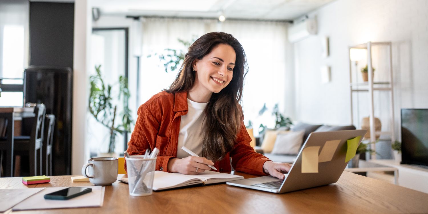 Eine Frau sitzt zuhause lächelnd am Tisch vor ihrem Laptop. Sie schreibt etwas in ein Notizbuch. . Artikel "Kompakte Fortbildungen zu Ernährungsbildung" öffnen.