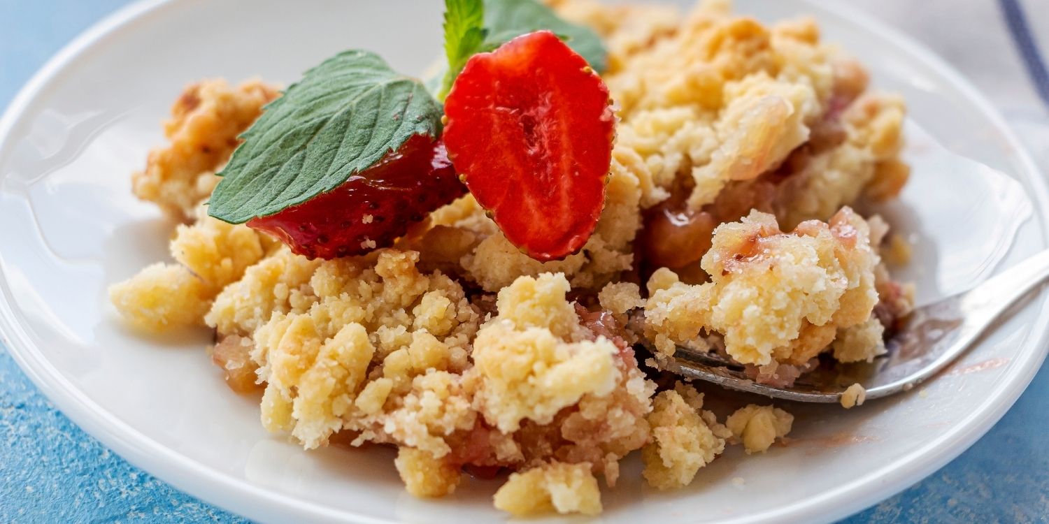 Auf einem hellblauen Untergrund steht ein weißer Teller mit Rhabarber-Crumble, dekoriert mit halbierten Erdbeeren und Melisse. . Artikel "Ideen für Desserts im Frühling" öffnen.