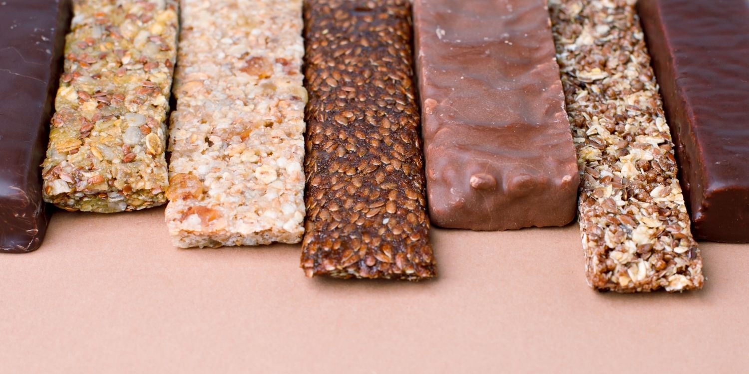 Aufgereiht nebeneinander liegen verschiedene Protein-, Schokoladen- und Müsliriegel am oberen Bildrand.. Artikel "Funktionelle Süßigkeiten im Trend" öffnen.