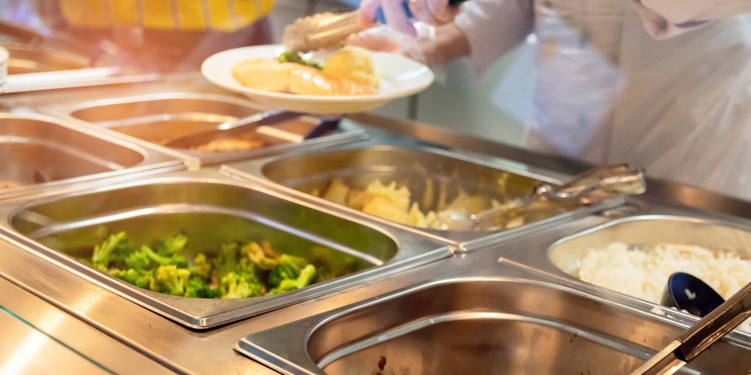 In einer Essensausgabe sind mehrere Edelstahlbehälter mit Brokkoli, Reis und Fisch gefüllt. Eine Küchenkraft steht dahinter und füllt einen Teller.. Artikel "Weniger Lebensmittelabfälle in der Gemeinschaftsverpflegung" öffnen.