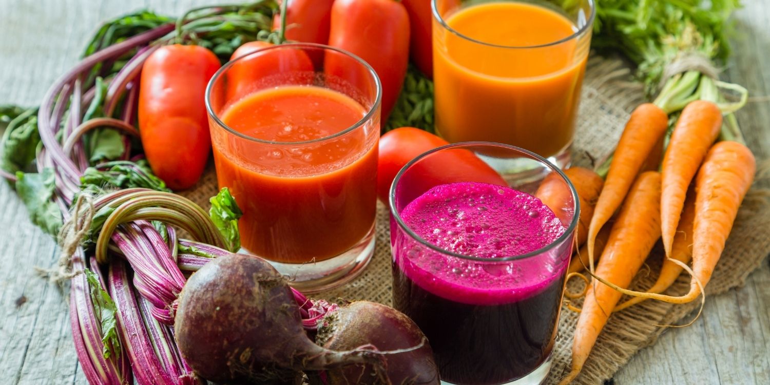 Auf einem Holztisch stehen drei Gläser mit Rote-Bete-, Möhren- und Tomatensaft. Daneben liegen Tomaten, Möhren und Rote-Bete-Knollen.. Artikel "Gemüsesaft im Trend" öffnen.