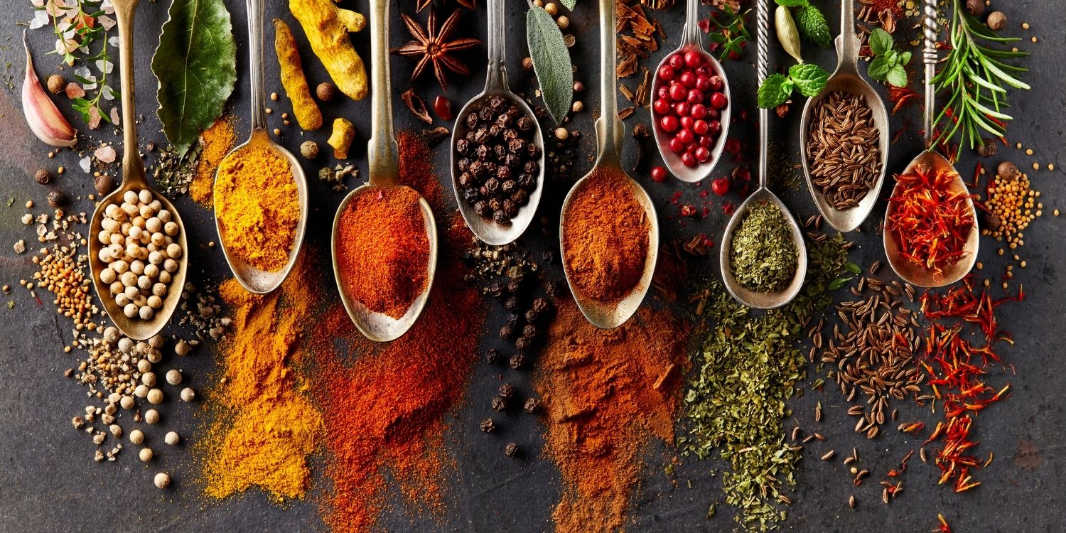 Auf einer dunklen Platte liegen neun Löffel nebeneinander mit verschiedenen Gewürzen darauf, etwa Pfefferkörner, Kurkuma, Paprika, Sternanis, Knoblauch und Rosmarin.. Artikel "Von A wie Anis bis Z wie Zimt" öffnen.