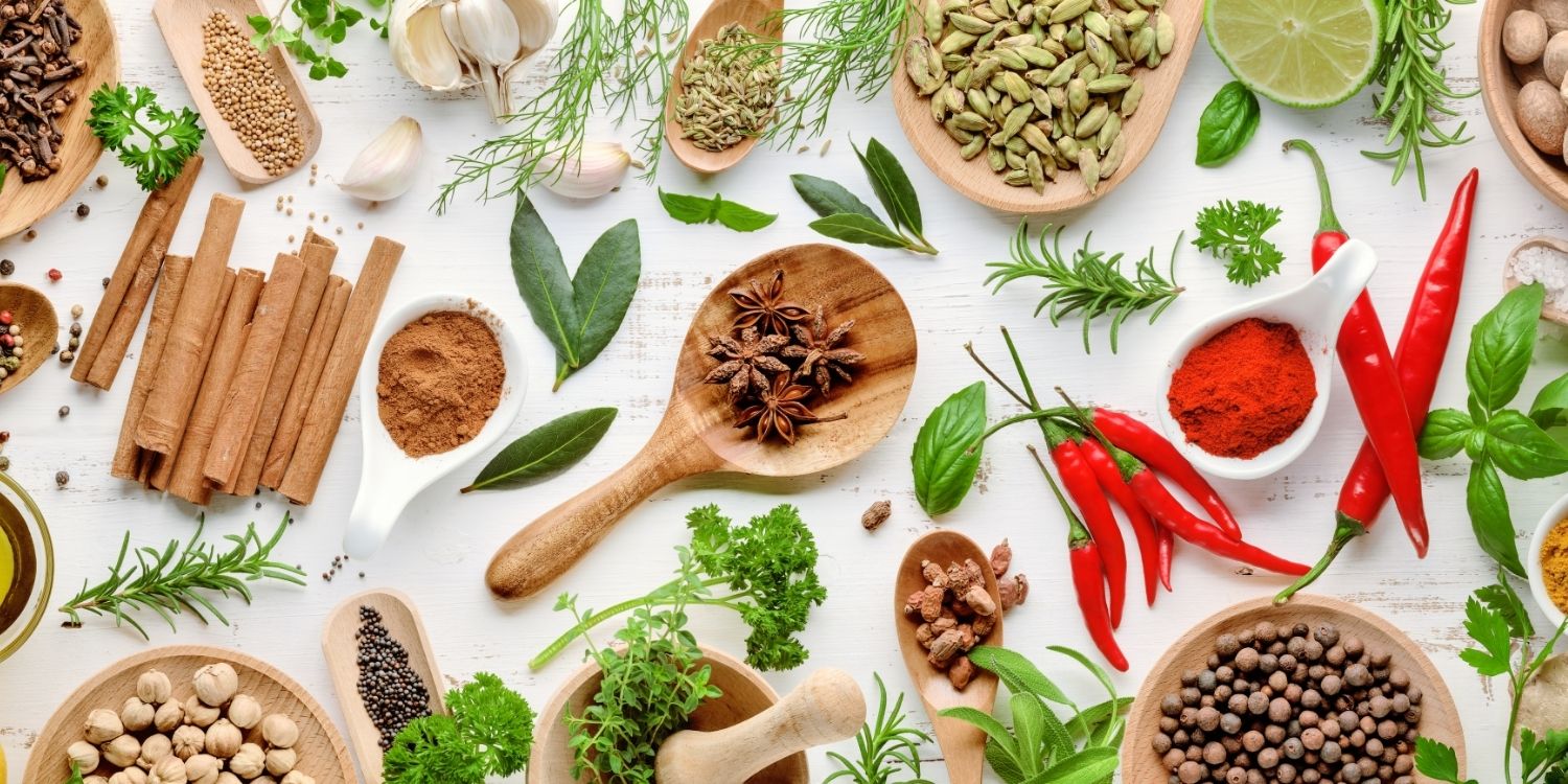 Auf einer weißen Fläche liegen verstreut frische Lorbeerblätter, Rosmarin, Basilikum, Dill und Petersilie. Außerdem Holzlöffel und Behälter mit Senfkörnern, Paprikapulver, Zimtstangen, Kardamom, Sternanis, Muskatnüssen, Peperoni und Knoblauchzehen.. Artikel "Die Welt der Gewürze und Kräuter" öffnen.