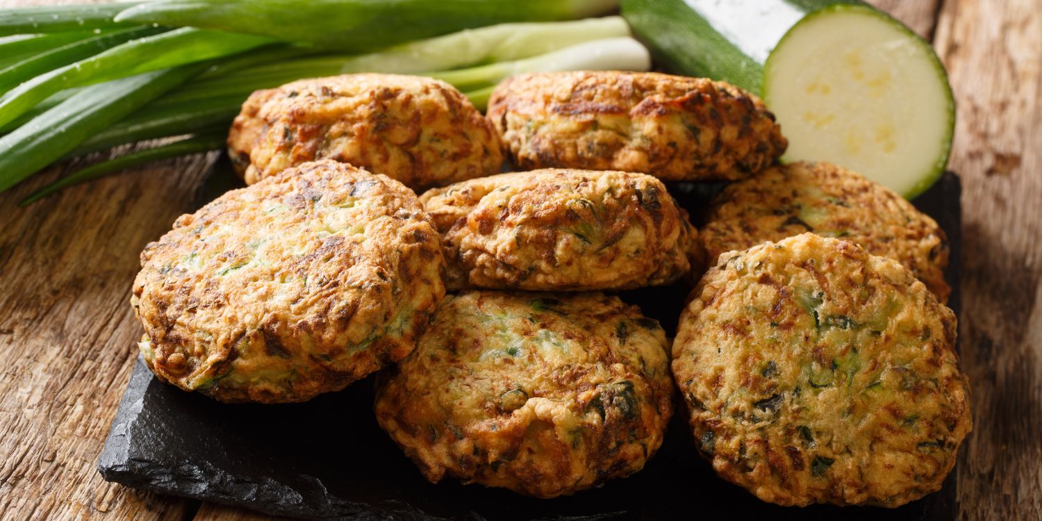 Auf einer schwarzen Schieferplatte liegen fertige Zucchinifrikadellen, daneben eine angeschnittene Zucchini und Frühlingslauch.. Artikel "Griechische Sommerküche" öffnen.