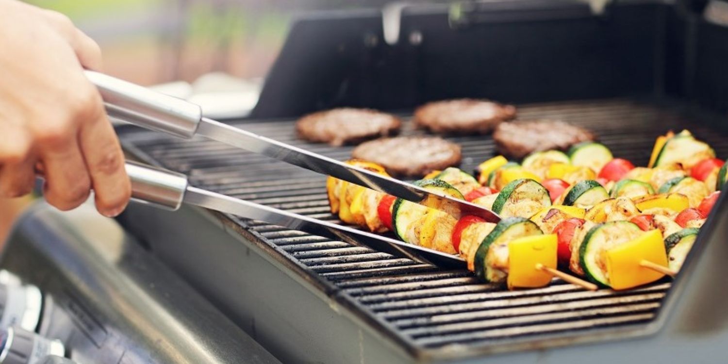 Auf einem Grillrost eines Gasgrills liegen Gemüsespieße und Fleisch-Patties. Ein Spieß wird mit einer Grillzange gewendet.. Artikel "Grillgenuss ohne Risiko" öffnen.