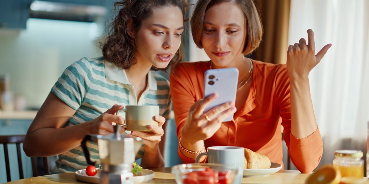 Zwei junge Frauen sitzen an einem gedeckten Frühstückstisch und schauen gemeinsam auf ein Handy.. Artikel "Wo und wie informieren sich junge Menschen über Ernährung?" öffnen.