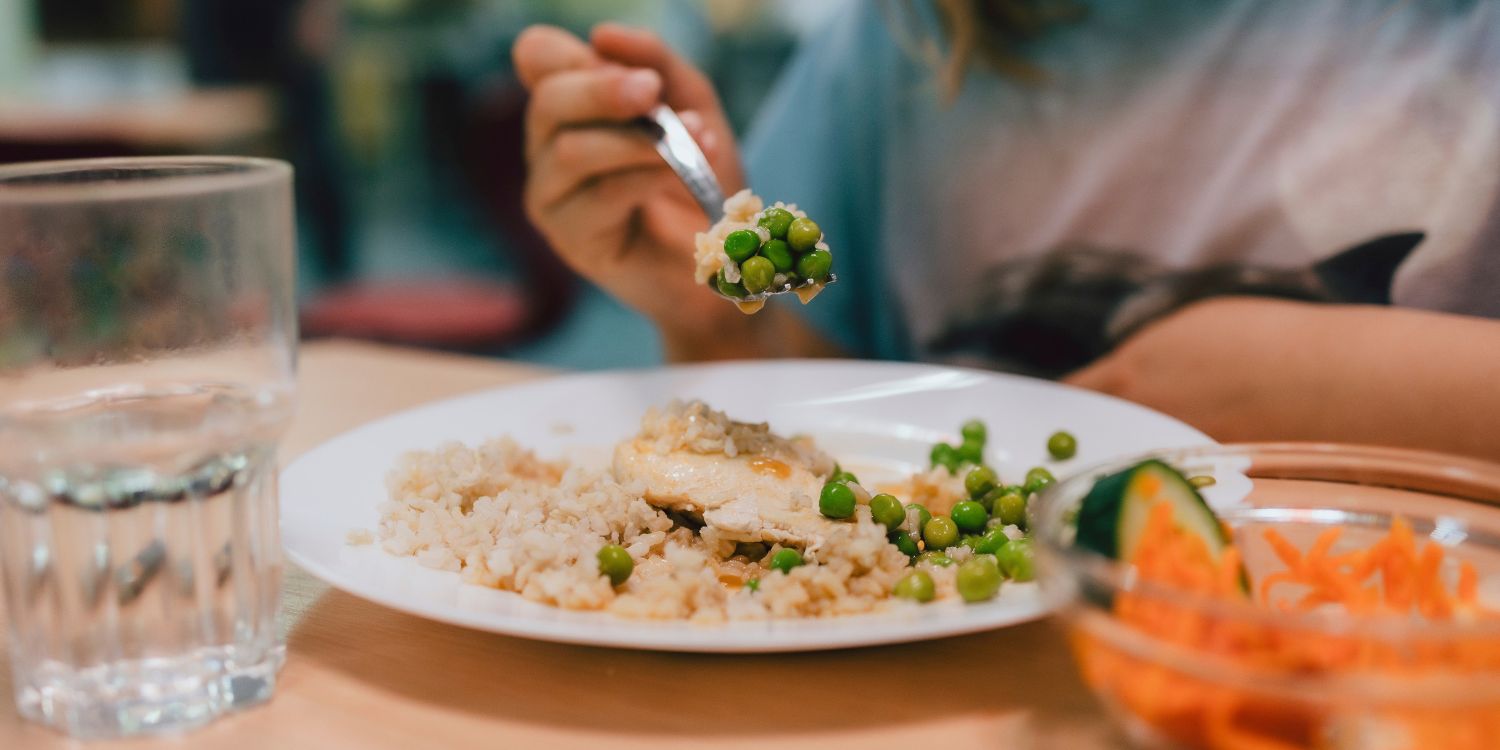 Ein Mädchen isst in einer Schulkantine Reis, Erbsen und Huhn von einem weißen Teller. Daneben steht ein Glas Wasser und ein Möhrensalat.. Artikel "Kita- und Schulverpflegung auf dem Prüfstand" öffnen.