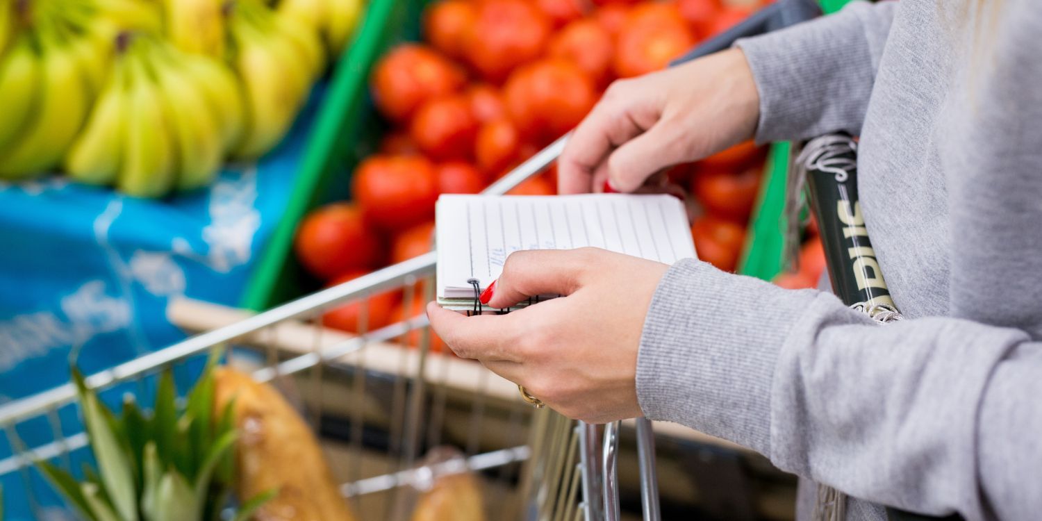 Eine Frau im grauen Sweatshirt schiebt einen Einkaufswagen durch die Gemüseabteilung und hält einen Block mit Notizen in der Hand.. Artikel "Kochen mit kleinem Budget" öffnen.