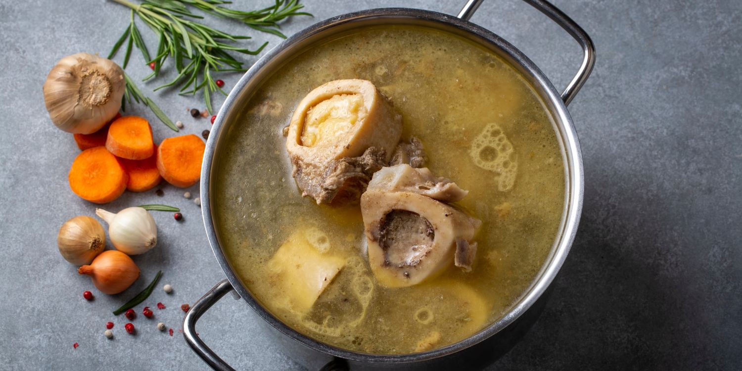 In einem Topf schwimmen Knochenstücke in einer Brühe. Neben dem Topf liegen Zwiebeln, Knoblauch, Möhren und Gewürze.. Artikel "Wirkt Knochenbrühe wirklich?" öffnen.