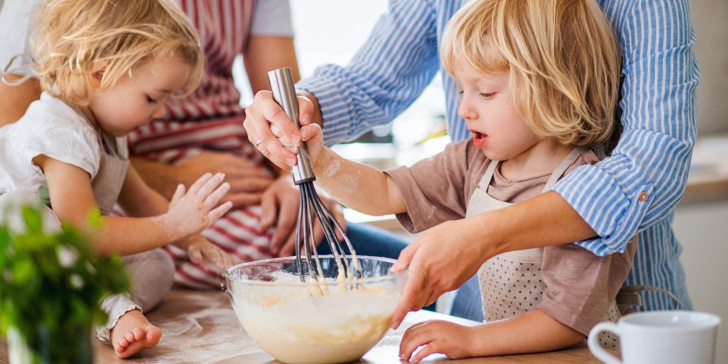In einer Küche führt eine Mutter die Hand eines blonden Kleinkindes beim Rühren von Teig. Ein anderes Kind sitzt mit bemehlten Händen auf der Arbeitsfläche beim Vater.. Artikel "Kochen mit Kindern " öffnen.